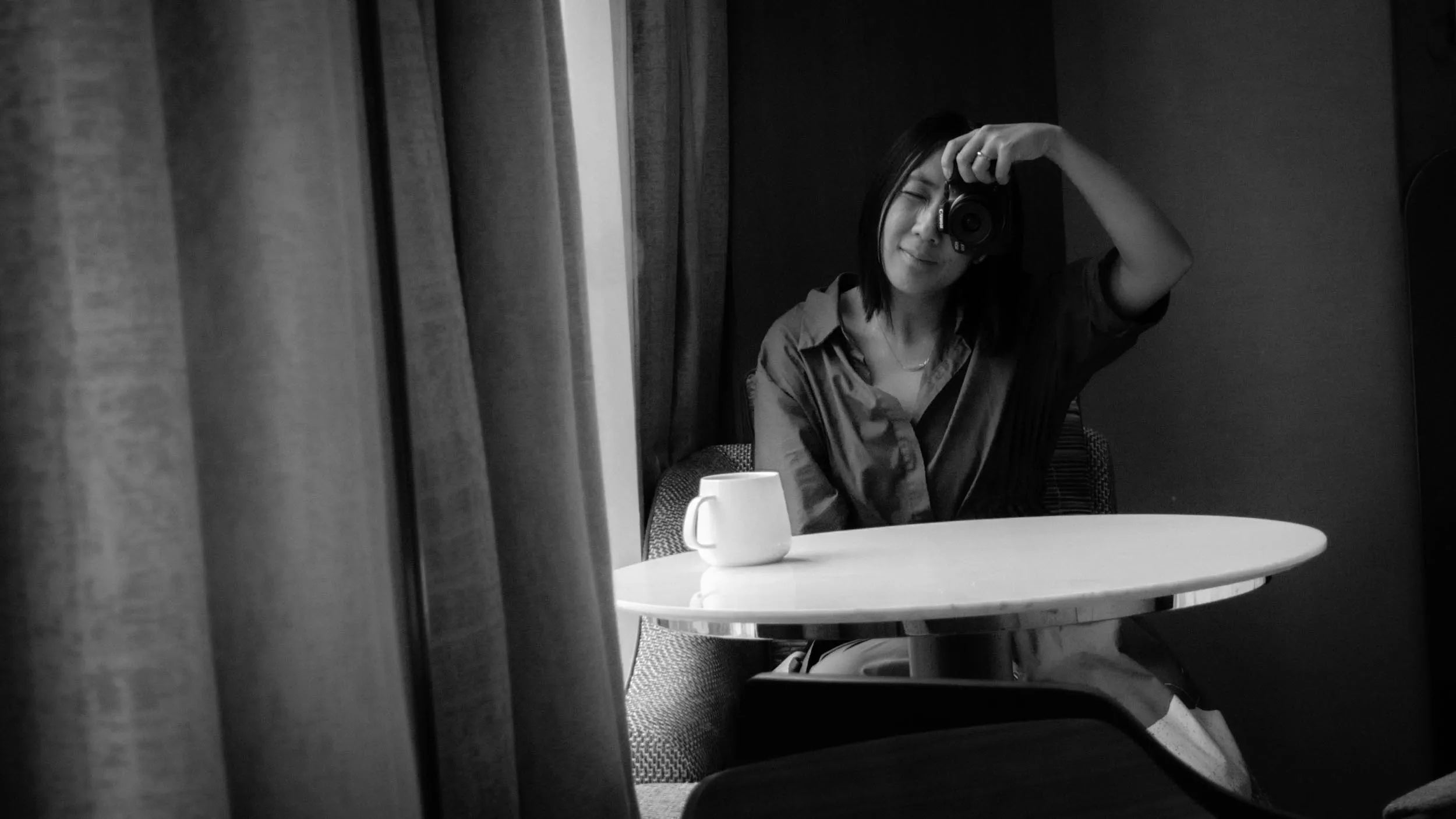 A black and white lifestyle photograph of a woman sitting at a small round table, holding a camera to her eye. A white mug sits on the table, with a large curtain framing the left side of the scene.