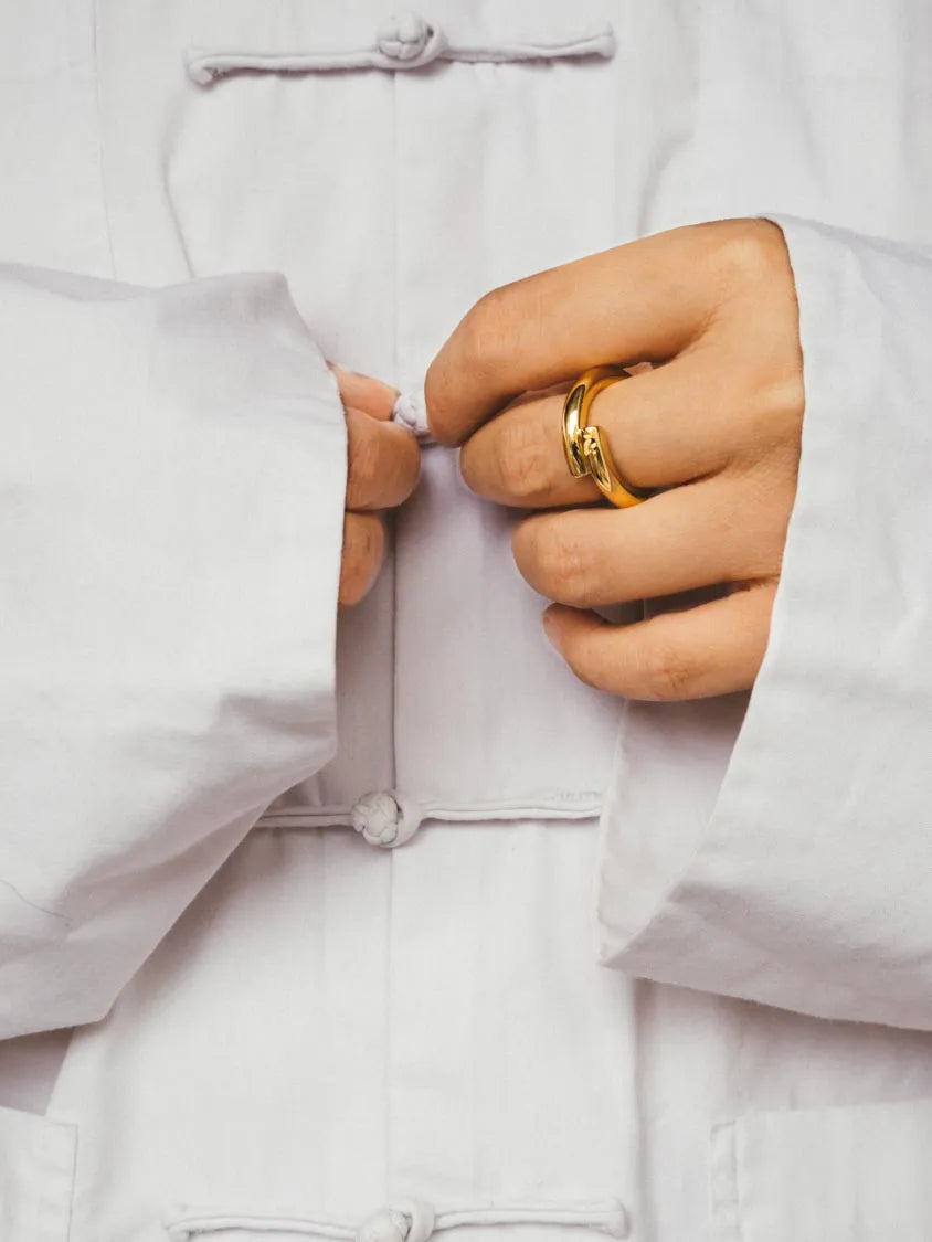 Close-up of a person's hands fastening a white traditional-style jacket with toggle pankou buttons, wearing a 18k gold ver,meil ring on one finger.
