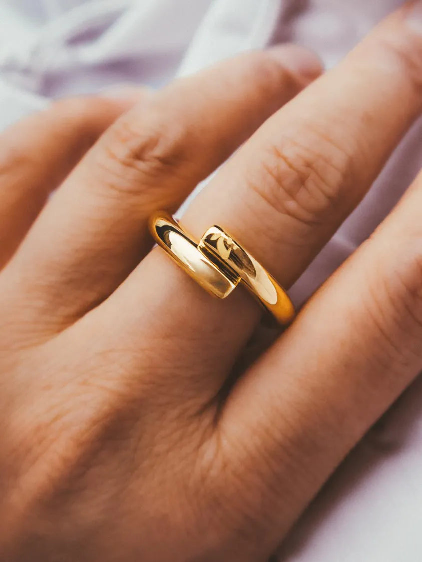 A close-up of a hand wearing a polished 18k gold vermeil ring with a unique interlocking band design, set against a soft, out-of-focus light background.